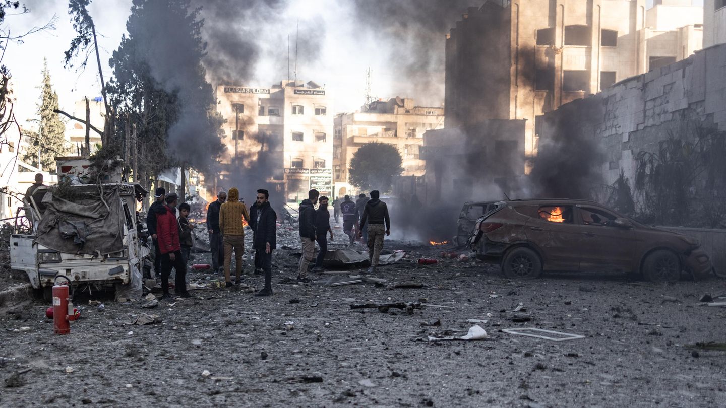 Eine Gruppe von Männern steht zwischen brennenden Fahrzeugen in einem zerbombten Wohnviertel. Eine Gruppe von Männern steht zwischen brennenden Fahrzeugen in einem zerbombten Wohnviertel.