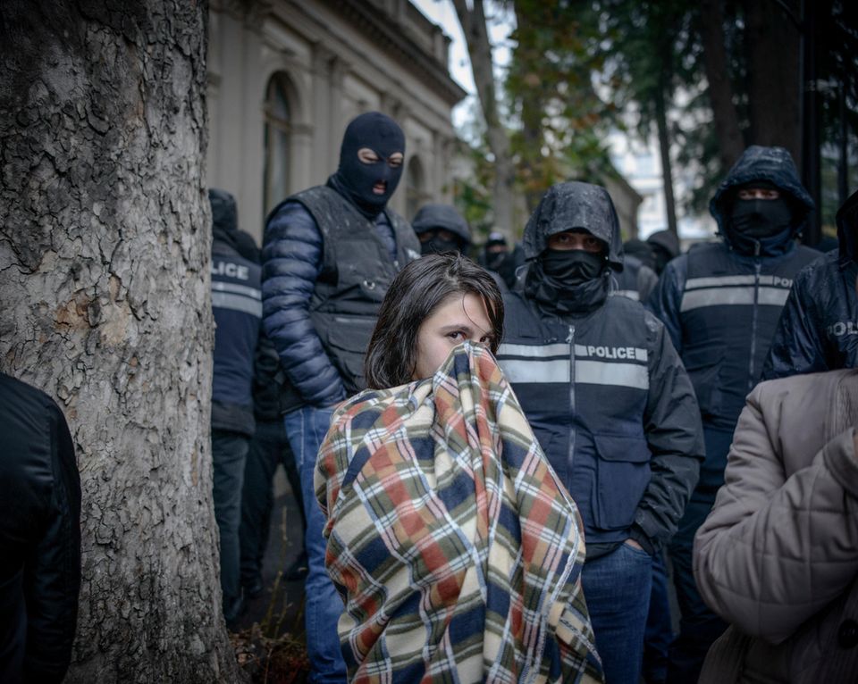 Frau in eine Decke eingewickelt. Hinter ihr vermummte Polizei