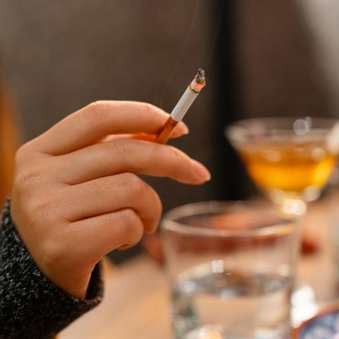 Eine Frauenhand hält eine angezündete Zigarette. Im Hintergrund sieht man verschwommen Gläser gefüllt mit Alkohol.
