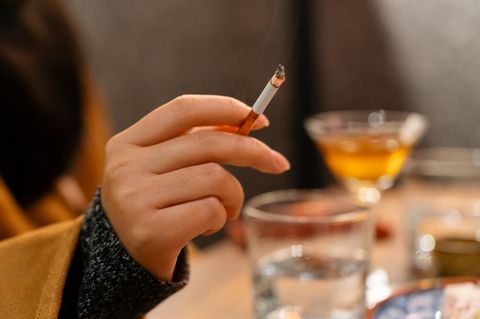 Eine Frauenhand hält eine angezündete Zigarette. Im Hintergrund sieht man verschwommen Gläser gefüllt mit Alkohol.