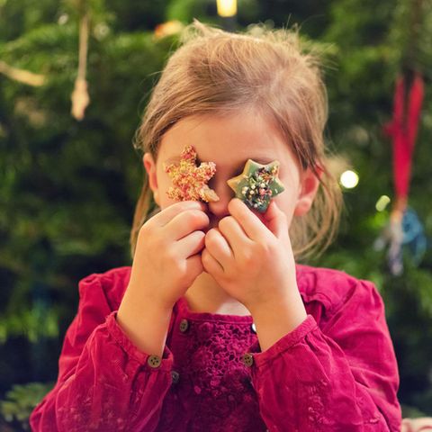 Ein Mädchen hält sich Kekse vor die Augen und steht vor einem Weihnachtsbaum