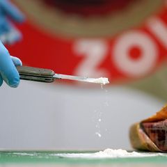 Ein geöffnetes Päckchen. Eine Hand in blauem Gummihandschuh hält ein Taschenmesser. Darauf liegt ein Häufchen weißes Pulver.