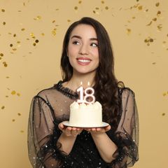 Frau hält köstlichen Kuchen mit zahlenförmigen Kerzen und schaut auf fallendes Konfetti vor beigem Hintergrund