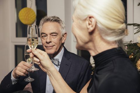 Ein Mann mit grauen Haaren und Sektglas in der Hand flirtet mit der Dame neben sich.
