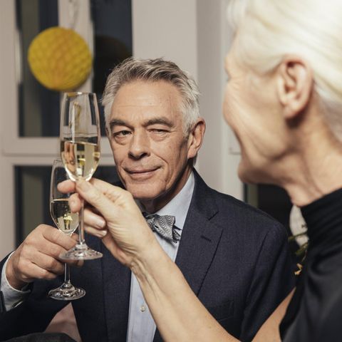 Ein Mann mit grauen Haaren und Sektglas in der Hand flirtet mit der Dame neben sich.