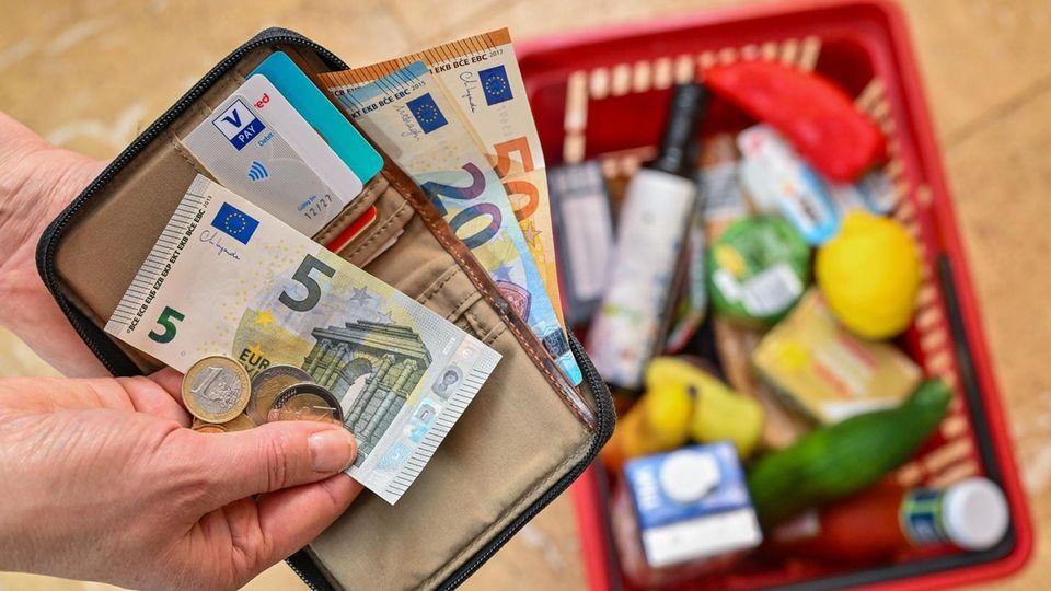 Eine Person hält ein Portemonnaie samt Geld in der Hand. Darunter befindet sich ein mit Lebensmitteln gefüllter Einkaufskorb