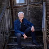 Robert Atzorn sitzt in blauer Jacke auf der Treppe eines Holzhäuschens in seinem Garten