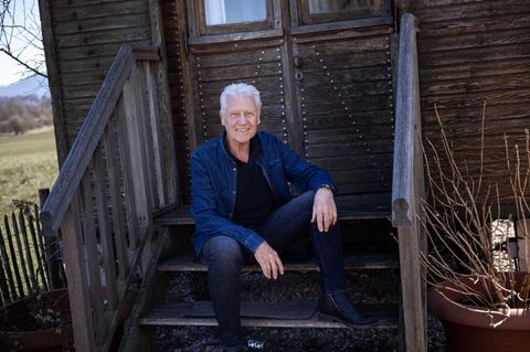 Robert Atzorn sitzt in blauer Jacke auf der Treppe eines Holzhäuschens in seinem Garten