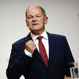 Olaf Scholz steht in schwarzem Anzug und roter Krawatte auf einer Bühne mit SPD-Logo und gestikuliert mit der rechten Hand