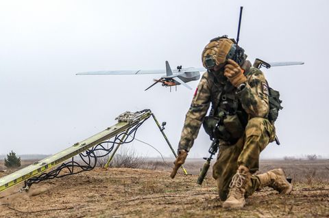 Ein russischer Soldat kniet auf einem Feld. Im Hintergrund startet ein unbemanntes Luftfahrzeug