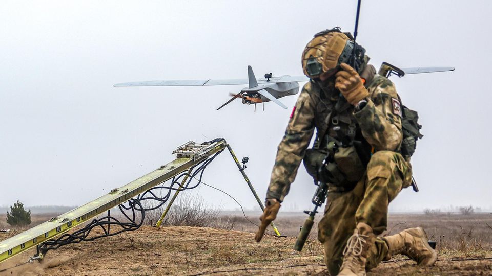 Ein russischer Soldat kniet auf einem Feld. Im Hintergrund startet ein unbemanntes Luftfahrzeug