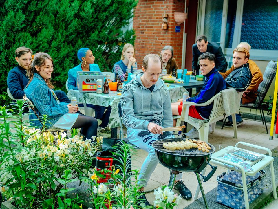 Sandmeyer in seiner Rolle als "Jonas Schulze" sitzt an einem kleinen Grill, im Hintergrund der restliche Cast Sandmeyer in seiner Rolle als "Jonas Schulze" sitzt an einem kleinen Grill, im Hintergrund der restliche Cast