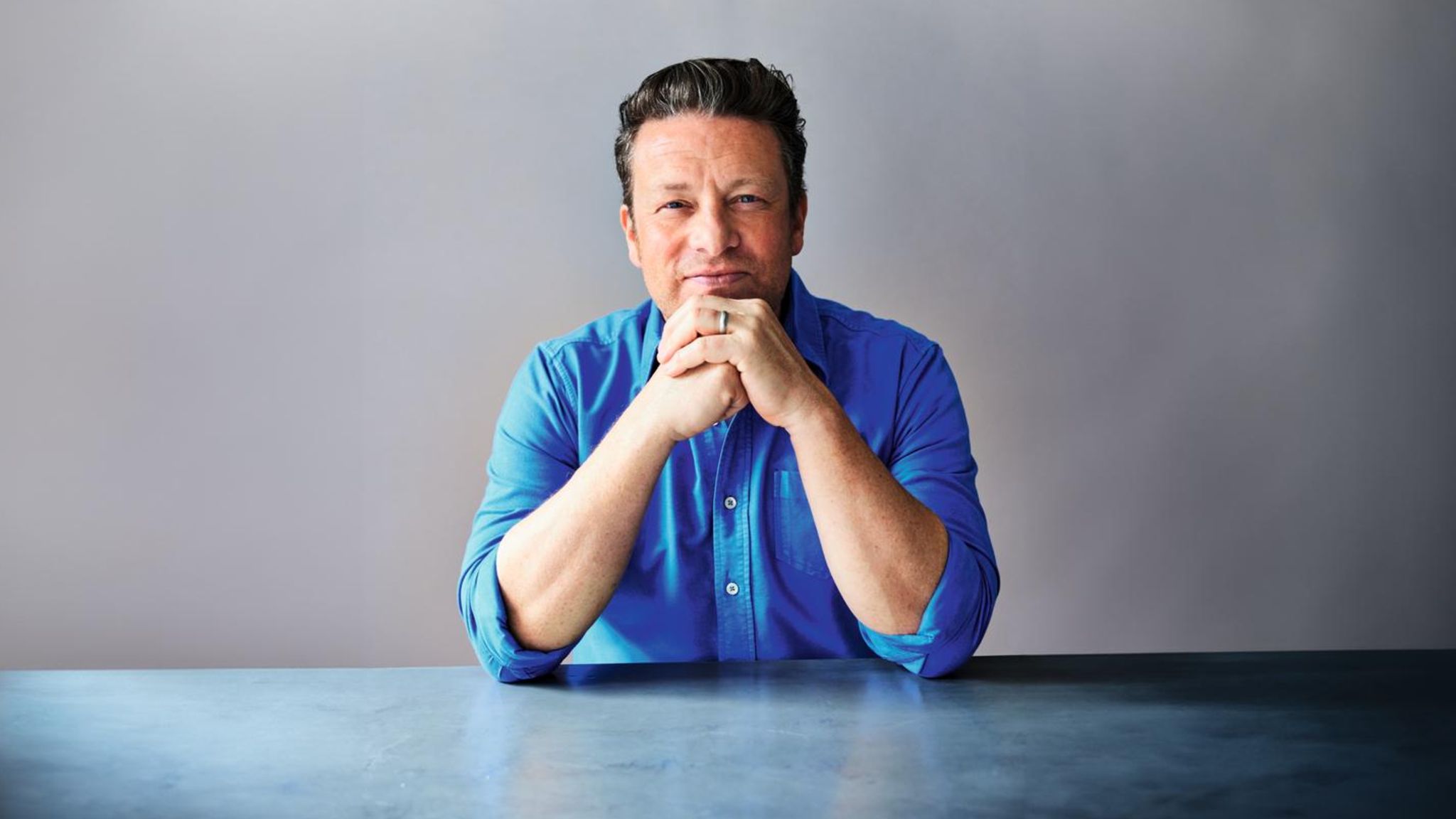 Jamie Oliver in einem blauen Hemd vor einer grauen Wand