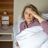 Erschöpfte Frau fasst sich im Bett liegend an den Kopf