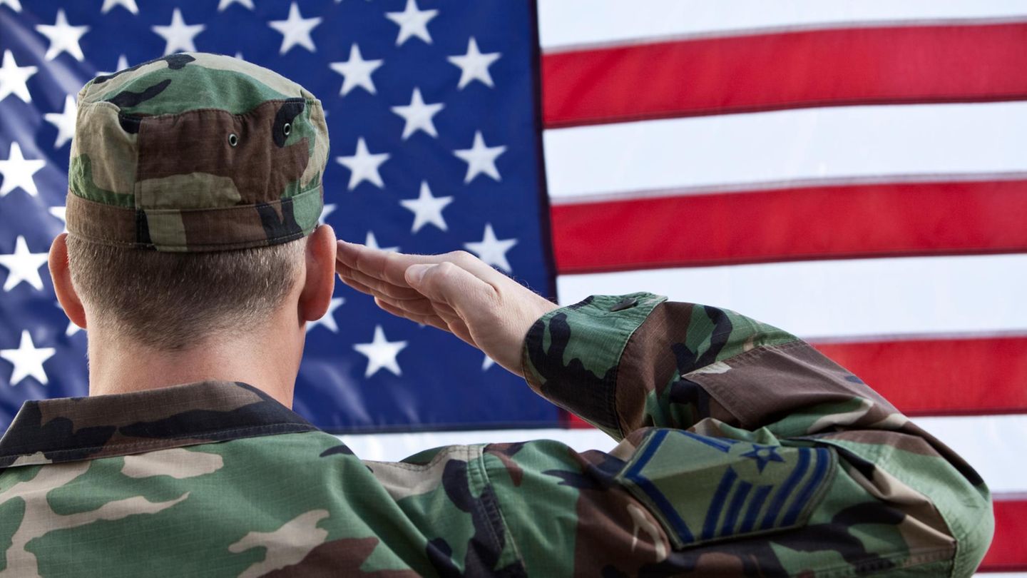 Ein Soldat in Tarn-Uniform salutiert vor einer Flagge der USA