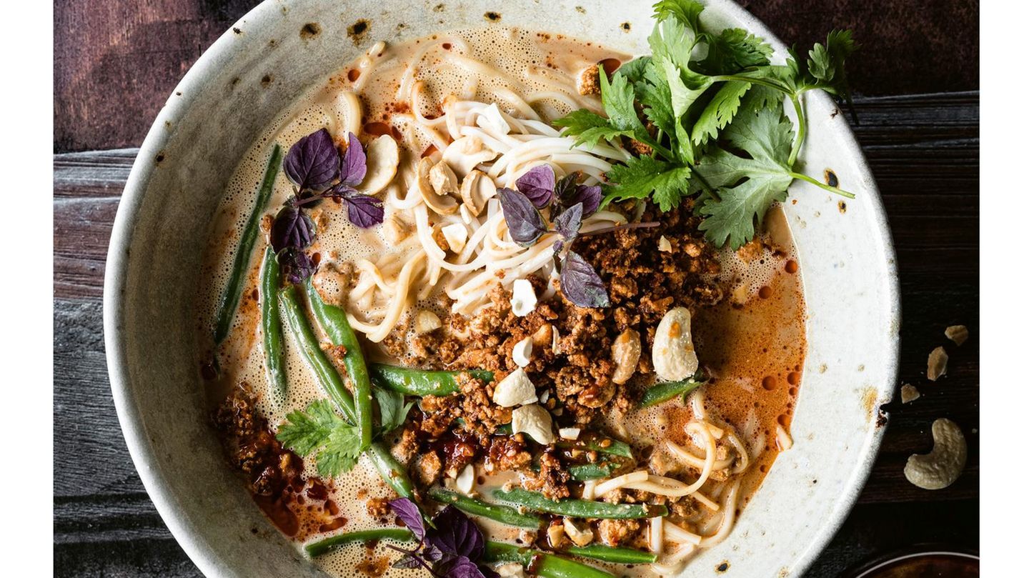    Zutaten für 4 Personen:  Für das Tofuhack  20 g Ingwer  2 Knoblauchzehen  250 g Tofu Natur  1 EL Pflanzenöl  1 EL geröstetes Sesamöl  2 EL Tomatenmark  4 EL dunkle Sojasauce     Für die Brühe  ca. 800 ml gute, kräftige Gemüsebrühe  500 ml Sojamilch  30-40 g helles, mildes Miso  100 g Tahin (Sesammus)  3-4 EL dunkle Sojasauce  2-3 TL Limettensaft  schwarzer Pfeffer aus der Mühle  Salz     Für Bohnen und Nudeln  300 g grüne Bohnen  200 g Ramennudeln  2-3 EL Cashewkerne Natur  1 EL Sriracha-Sauce  1 EL Pflanzenöl  2 EL Korianderblätter  1 EL rote Shiso (asiatische Kresse)  Salz     Zubereitung  1. Für das Tofuhack Ingwer und Knoblauchzehen schälen und fein hacken. Den Tofu gut trocken tupfen, dann fein zerkrümeln. Pflanzenöl und Sesamöl in einer beschichteten Pfanne erhitzen und die Tofukrümel darin zwei bis drei Minuten kräftig anbraten. Ingwer und Knoblauch dazugeben und kurz mit anbraten. Tomatenmark und Sojasauce unterrühren, abschmecken und die Pfanne beiseitestellen.      2. Für die Brühe Gemüsebrühe, Sojamilch, Miso, Tahin und drei Esslöffel Sojasauce in einen Topf geben und bei mittlerer Hitze etwa fünf Minuten offen einkochen lassen, dann mit restlicher Sojasauce, Limettensaft, etwas Salz und Pfeffer abschmecken und vom Herd nehmen. Falls die Brühe krisselig wird (gerinnt), mit dem Stabmixer kurz pürieren, dann wird sie wieder glatt und cremig.      3. Inzwischen die Bohnen putzen, waschen, in kochendem Salzwasser sieben bis zehn Minuten blanchieren, dann in ein Sieb abgießen, kalt abbrausen und abtropfen lassen.     4. Währenddessen die Nudeln nach Packungsangaben kochen, in ein Sieb abgießen, kalt abbrausen und abtropfen lassen.      5. Cashewkerne grob hacken, in einer Pfanne ohne Fett kurz anrösten und beiseitestellen. Sriracha-Sauce und Öl verrühren.      6. Die Brühe erhitzen. Das Tofuhack in der Pfanne unter Rühren noch mal heiß werden lassen.      7. Tofuhack, Nudeln und Bohnen in Schalen geben, mit der sehr heißen Brühe begießen, das Sriracha-Öl hineinträufeln und mit Cashews, Koriander und Shiso bestreuen. 