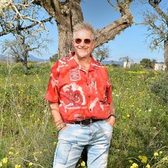 Martin Semmelrogge im roten Hemd auf seiner Finca, im Hintergrund blauer Himmel