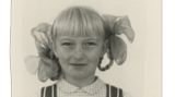 Ein Porträt der 9-jährigen Irina. Sie trägt ein schickes Kleid, die Haare mit Schleifen darin sind zu Zöpfen geflochten
