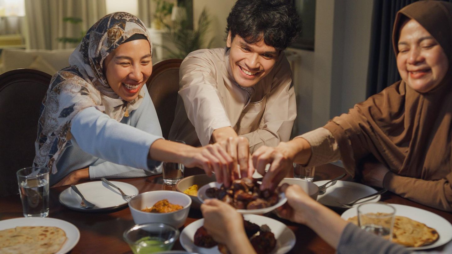 Fastenbrechen im Ramadan: eine dreiköpfige muslimische Familie sitzt am Tisch und greift lächelnd nach Essen