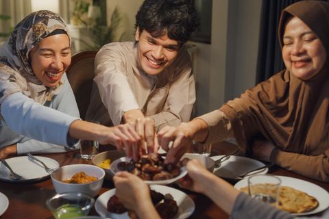 Fastenbrechen im Ramadan: eine dreiköpfige muslimische Familie sitzt am Tisch und greift lächelnd nach Essen