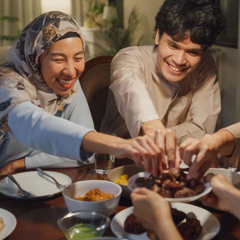 Fastenbrechen im Ramadan: eine dreiköpfige muslimische Familie sitzt am Tisch und greift lächelnd nach Essen