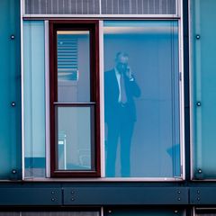 Friedrich Merz steht telefonierend hinter einer Glasscheibe