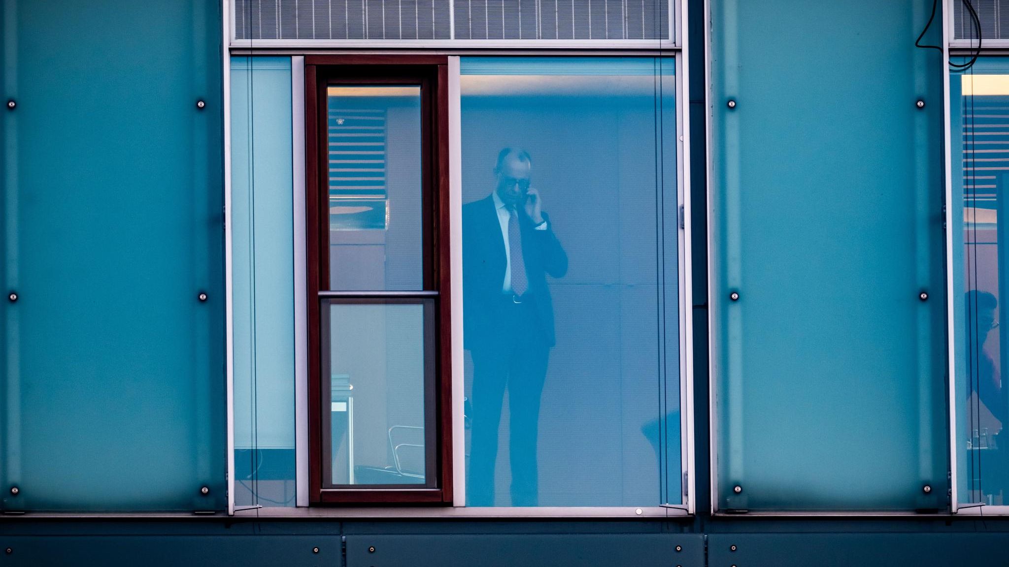 Friedrich Merz steht telefonierend hinter einer Glasscheibe