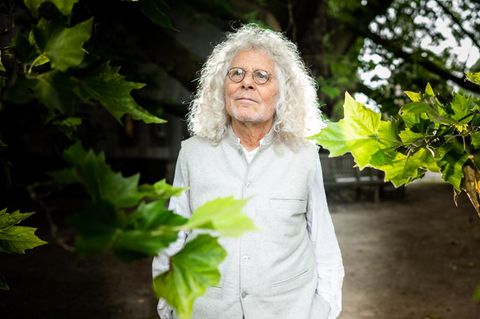 Rainer Langhans steht in weißen Klamotten zwischen Blättern eines Ahornbaumes und blickt in die Ferne