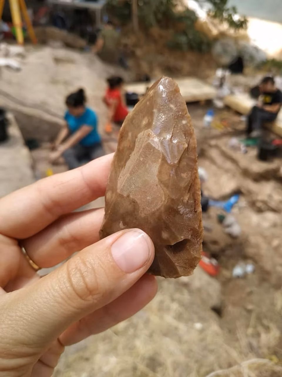 Eine Hand hält ein mehrere Jahrzehntausende altes Steinwerkzeug, dahinter Archäologen bei der Arbeit Eine Hand hält ein mehrere Jahrzehntausende altes Steinwerkzeug, dahinter Archäologen bei der Arbeit