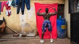 Bodybuilder Tamale Safalu steht vor einer Wand und hebt die Arme nach oben.
