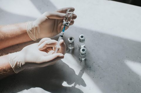 Eine Hand mit Latexhandschuhen hält ein Fläschchen mit einer klaren Flüssigkeit und eine Spritze. Thema Pflege