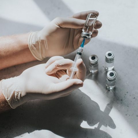 Eine Hand mit Latexhandschuhen hält ein Fläschchen mit einer klaren Flüssigkeit und eine Spritze. Thema Pflege