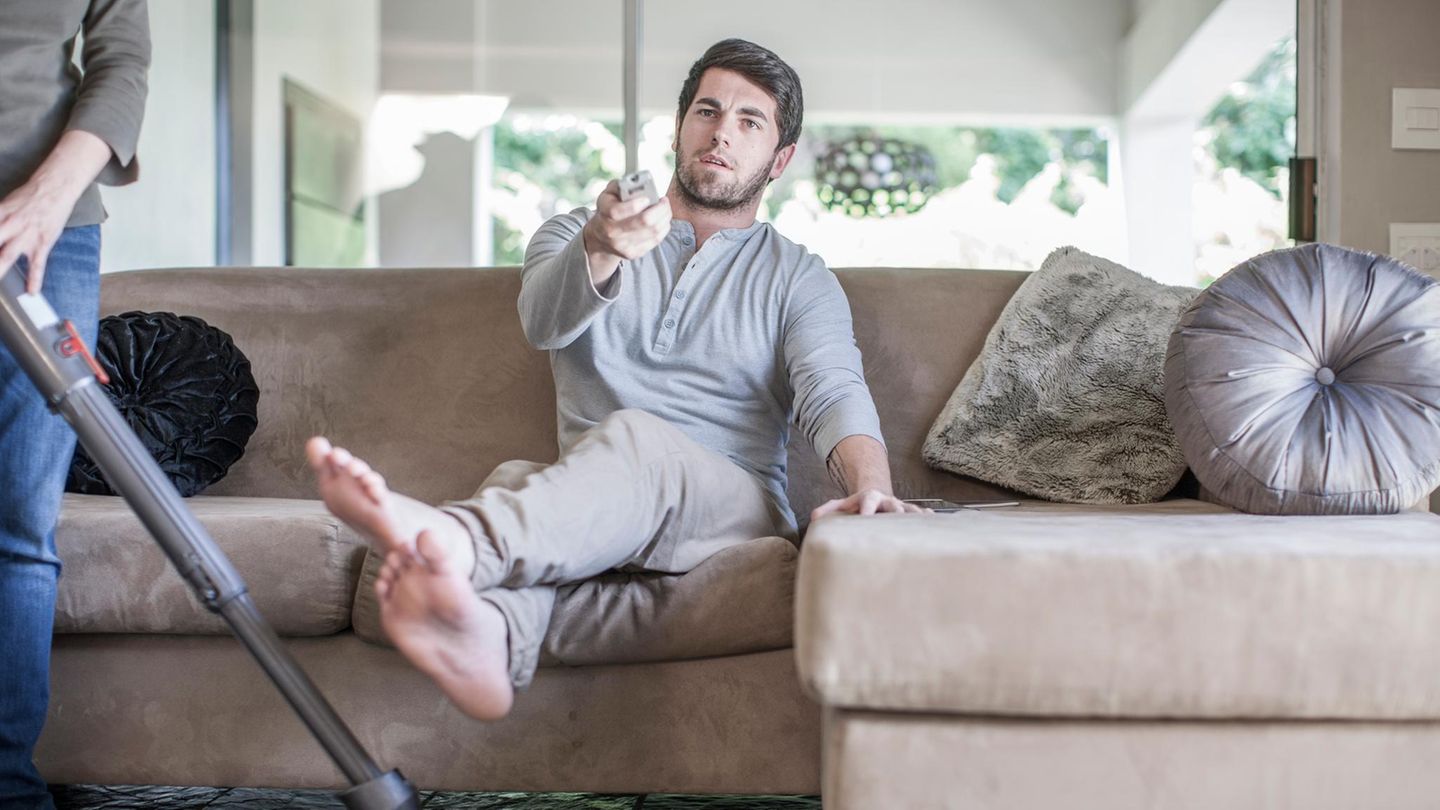 Ein Mann sitzt auf der Couch und schaut TV, während die Frau den Haushalt macht