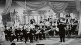 Als Mitglied der Europäischen Rundfunkunion (EBU) überträgt die ARD seit 1956 den Eurovision Song Contest, vormals Grand Prix Eurovision de la Chanson. Gleich im zweiten Jahr fand der Wettbewerb in Frankfurt am Main statt. Das Foto zeigt die Tanzorchester des Hessischen Rundfunks unter Leitung von Willy Berking (vorn stehend) im Sendesaal des Hessischen Rundfunks. Deutschland gewann den Wettbewerb bislang zweimal, 1982 mit Nicole und 2010 mit Lena Meyer-Landrut