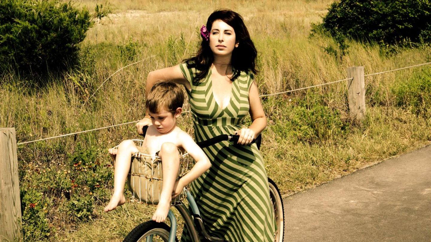 Mütterrente. Frau im Retro-Look transportiert ihren Sohn im Fahrradkorb Mütterrente. Frau im Retro-Look transportiert ihren Sohn im Fahrradkorb