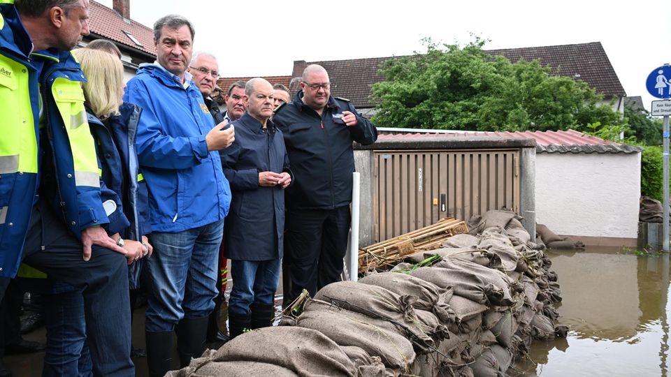 Markus Söder und Olaf Scholz stehen in Gummistiefeln hinter Sandsäcken vor den Starkregen-Fluten.