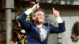 In einen blauen Anzug gekleideter Mann im Luftsprung, die beiden Zeigefinger in die Luft gestreckt