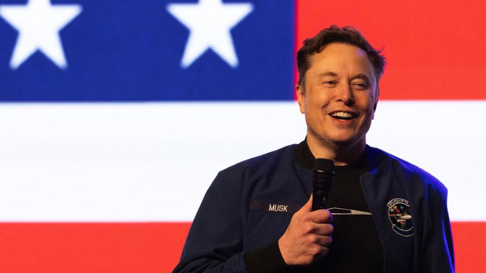 Elon Musk hält auf einer Bühne eine Rede. Im Hintergrund ist die US-amerikanische Nationalflagge