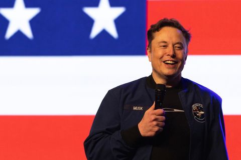 Elon Musk hält auf einer Bühne eine Rede. Im Hintergrund ist die US-amerikanische Nationalflagge
