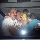 Rudi Völler und Lothar Matthäus sitzen im Mannschaftsbus und halten den WM-Pokal fest