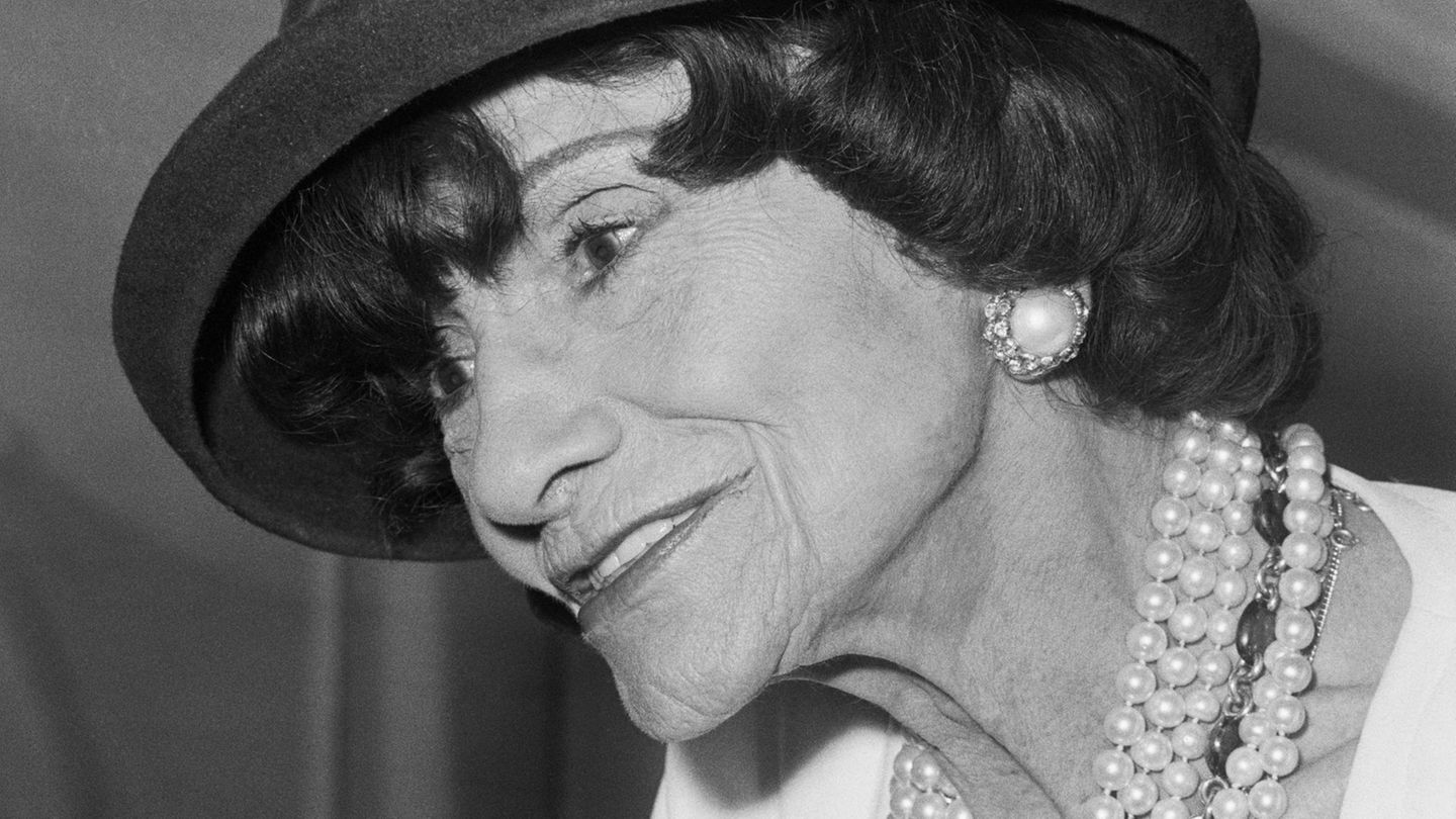 Im Portrait: Coco Chanel mit schwarzem Hut, weißen Perlenohrringen und Perlenketten
