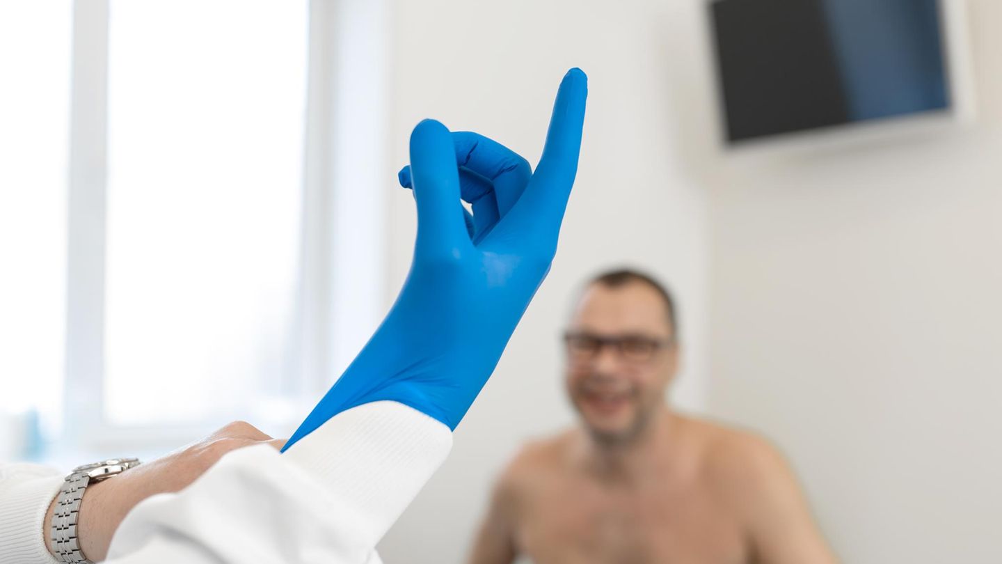 Prostatakrebs-Untersuchung beim Arzt: Ein Arzt zieht sich einen blauen Gummihandschuh an, ein Patient sitzt im Hinergrund