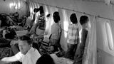 Kinder an Bord eines Flugzeugs stehen an den Fenstern und schauen nach draußen. Am Boden liegen weitere Kinder und schlafen