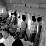 Kinder an Bord eines Flugzeugs stehen an den Fenstern und schauen nach draußen. Am Boden liegen weitere Kinder und schlafen