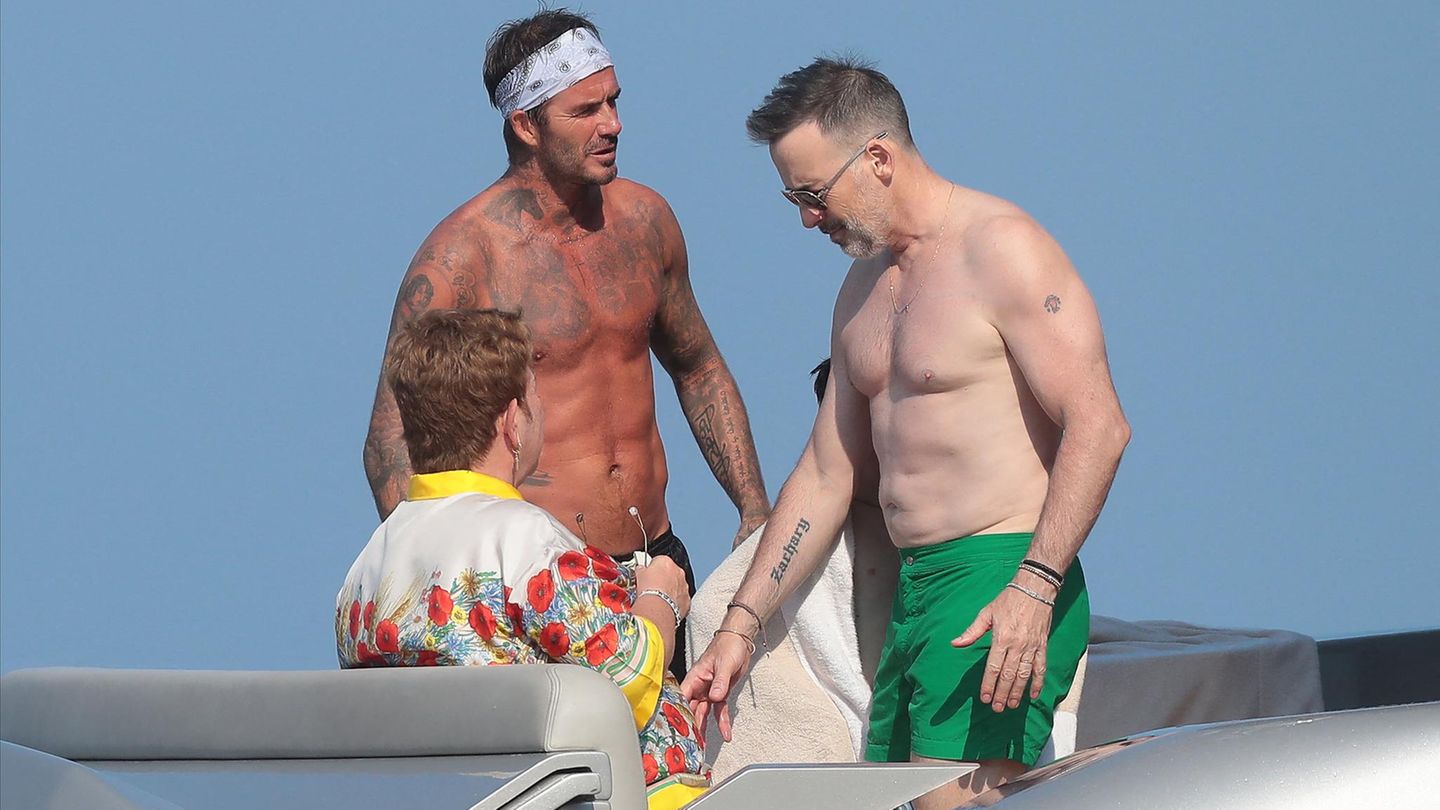 David Beckham mit nacktem Oberkörper und Stirnband auf einer Yacht, links und rechts von ihm Elton John und Mann