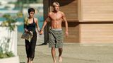 David Beckham in langer Badehose Hand in Hand mit Ehefrau Victoria im schwarzen Kleid am Strand