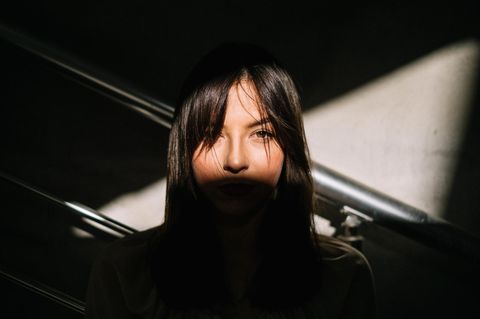 Gesicht einer jungen Frau in einem Streifen Sonnenlicht. Sie hat dunkle Haare und möglicherweise Selbstzweifel