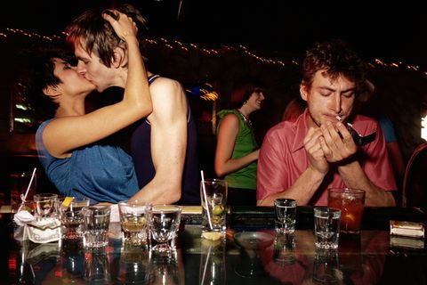 An einer Bar küsst sich ein Paar während ein Mann daneben eine Zigarette raucht