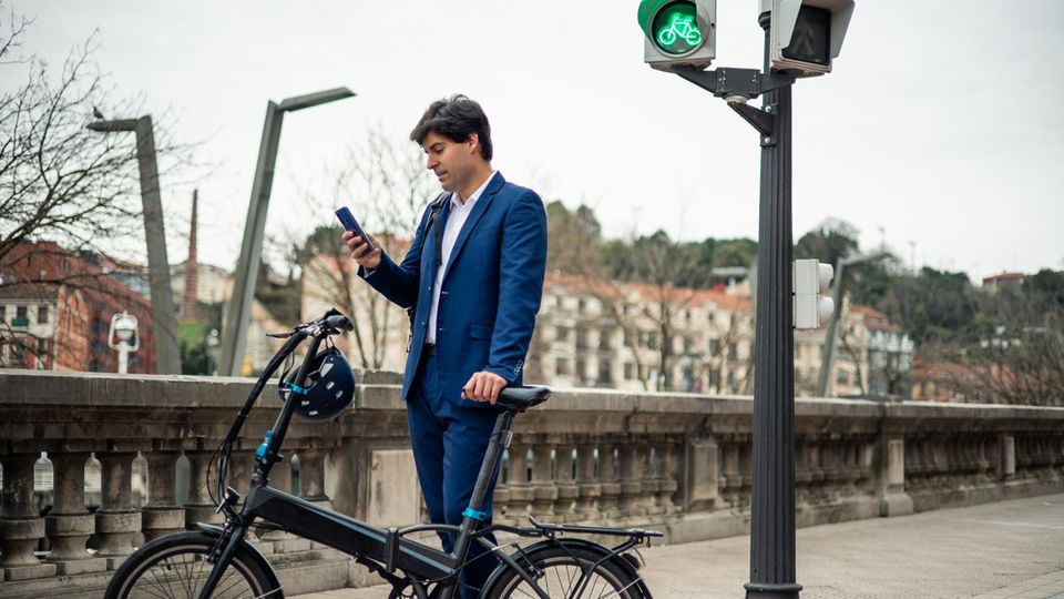 Mann im Anzug steht neben einer Fahrradampel mit einem Pendler-E-Bike. Er schaut aufs Handy Mann im Anzug steht neben einer Fahrradampel mit einem Pendler-E-Bike. Er schaut aufs Handy