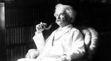 Mark Twain mit Pfeife und weißem Anzug in einem Sessel.